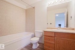 Bathroom featuring a white bathtub with beige tiled surround, a toilet, and a wood-finish vanity with an integrated sink and light-colored countertop - 