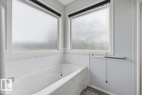 White soaking tub with surrounding white ceramic tile, framed by two privacy glass windows with black roller shades - 3570 Mclean Crescent, Edmonton, AB - Indoor Photo Showing Bathroom