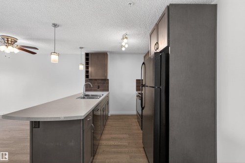 Kitchen featuring a spacious island with an integrated sink, dark wood-finish cabinetry, a subway tile backsplash, and black appliances - 215 6084 Stanton Drive, Edmonton, AB - Indoor Photo Showing Kitchen With Double Sink