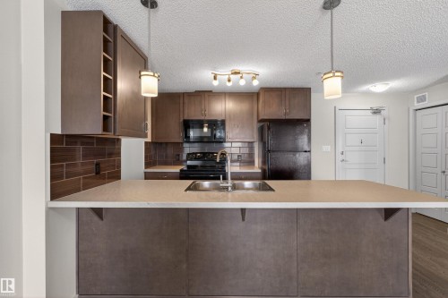 Kitchen featuring a breakfast bar with a light-toned countertop, wood-finish cabinetry, stainless steel double-basin sink, and dark subway tile backsplash - 215 6084 Stanton Drive, Edmonton, AB - Indoor Photo Showing Kitchen With Double Sink