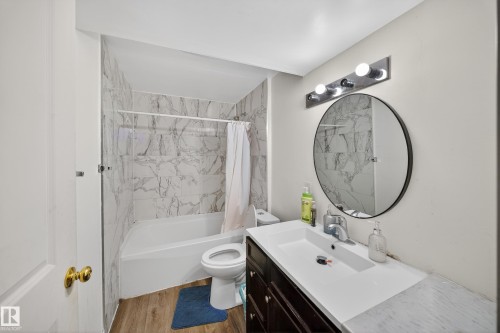 This bathroom features a full-sized bathtub with white and grey marble-style tiled walls, a dark wood vanity with a white countertop and integrated sink, and a circular mirror with a modern light fixture overhead - 3517 25 Street, Edmonton, AB - Indoor Photo Showing Bathroom