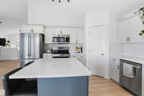 Kitchen featuring a central island with a white countertop and blue-painted base, stainless steel appliances, white cabinetry with gold-toned hardware, light wood-finish flooring, and vaulted ceilings - 396 Kananaskis Way, Devon, AB - Indoor Photo Showing Kitchen