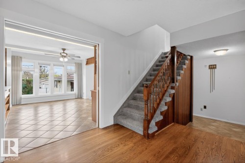 Foyer with wood-finish flooring opening to a carpeted staircase with wood banister and a separate room featuring large windows, tiled flooring, and a ceiling fan - 8911 84 Avenue, Edmonton, AB - Indoor