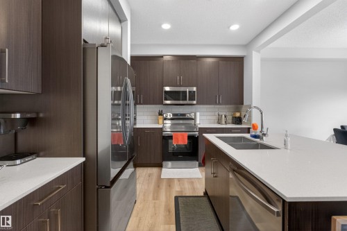 Modern kitchen featuring dark wood-finish cabinetry, stainless steel appliances, light-toned countertops, a subway tile backsplash, and wood-finish flooring - 332 Juniper Cove, Leduc, AB - Indoor Photo Showing Kitchen With Double Sink