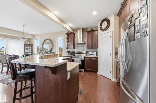 Kitchen featuring a breakfast bar with granite-finish countertop, dark wood-finish cabinetry, stainless steel appliances, and wood-finish flooring - 1590 Chapman Way, Edmonton, AB 