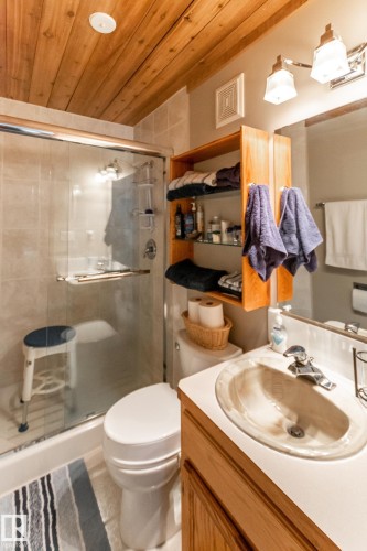 Bathroom featuring a wood-panel ceiling, tiled shower with glass enclosure, single vanity with integrated sink, wood-finish cabinetry, and wall-mounted light fixture - 4611 Lakeshore Drive, Bonnyville Town, AB - Indoor Photo Showing Bathroom