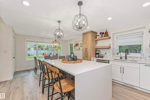 Contemporary kitchen featuring a large waterfall island with seating, white shaker-style cabinetry, wood-finish flooring, geometric pendant lighting, and a patterned backsplash - 627 Forest Lane, Edmonton, AB - Indoor