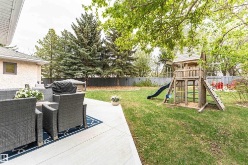 Spacious backyard featuring a large concrete patio, mature trees providing natural shade, a substantial wood-built play structure with a slide, and a durable metal fence enclosing the perimeter - 627 Forest Lane, Edmonton, AB - Outdoor