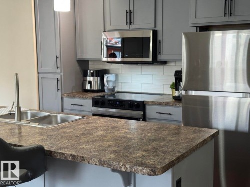 Kitchen featuring an island with double stainless-steel sinks, grey cabinetry, stainless-steel appliances, and a white subway tile backsplash - 228 308 Ambleside Link Sw, Edmonton, AB - Indoor Photo Showing Kitchen With Double Sink With Upgraded Kitchen