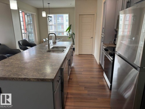 Kitchen featuring a double basin stainless steel sink, wood-finish flooring, stainless steel appliances, grey cabinetry, and a stone-finish countertop - 228 308 Ambleside Link Sw, Edmonton, AB - Indoor Photo Showing Kitchen With Double Sink