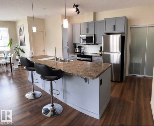 Kitchen island with a dual-basin stainless steel sink, light gray cabinetry, stainless steel appliances, and wood-finish flooring - 228 308 Ambleside Link Sw, Edmonton, AB - Indoor Photo Showing Kitchen With Double Sink With Upgraded Kitchen