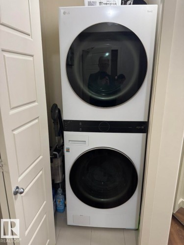 Stacked white LG washer and dryer unit, front-loading design, featuring dark glass doors and integrated controls, set within a compact utility alcove - 228 308 Ambleside Link Sw, Edmonton, AB - Indoor Photo Showing Laundry Room