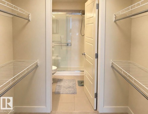 Walk-in closet with wire shelving and hanging rods, opening to a bathroom featuring a glass-enclosed shower and tiled flooring - 228 308 Ambleside Link Sw, Edmonton, AB - Indoor With Storage