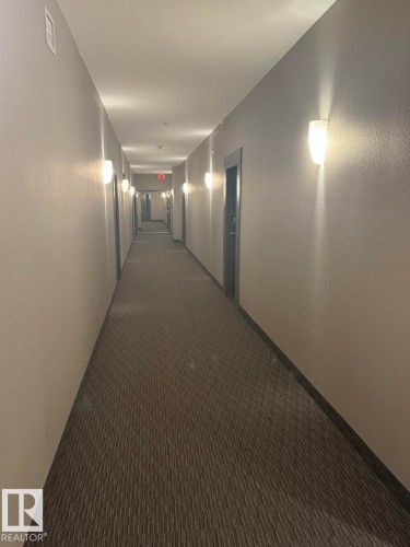 Hallway featuring recessed ceiling lighting, wall-mounted sconces, neutral wall finishes, patterned carpet flooring, and dark trim around doorways - 228 308 Ambleside Link Sw, Edmonton, AB - Indoor Photo Showing Other Room