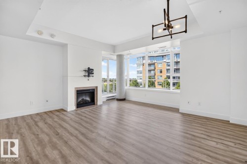 Living area featuring wood-finish flooring, a contemporary fireplace with a light-toned surround, expansive windows, and a modern chandelier - #407 2504 109 St Nw, Edmonton, AB - Indoor Photo Showing Living Room With Fireplace