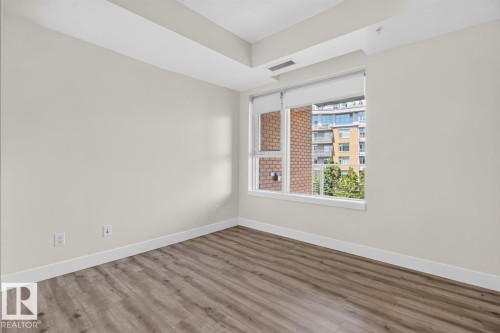 Room featuring wood-finish flooring, neutral wall paint, white baseboards, and a large window with roller shades - #407 2504 109 St Nw, Edmonton, AB - Indoor Photo Showing Other Room