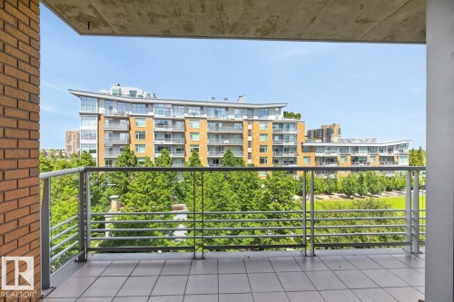 Balcony featuring tile flooring and metal railings, overlooking a tree-lined green space with urban buildings in the background - #407 2504 109 St Nw, Edmonton, AB - Outdoor With Balcony