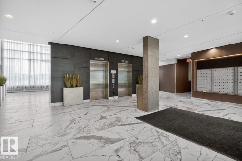 Modern building lobby featuring polished marble flooring, dark textured wall panels, a concrete pillar, and stainless steel elevators - #407 2504 109 St Nw, Edmonton, AB - Indoor Photo Showing Other Room