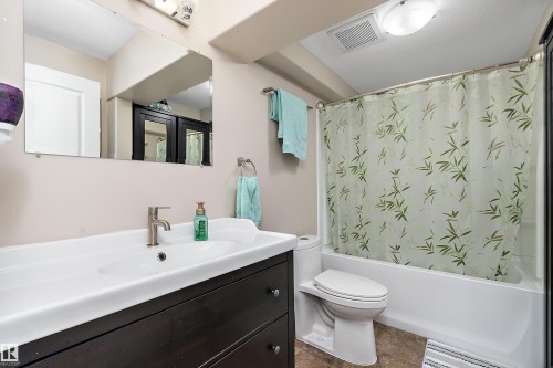 Bathroom featuring a vanity with an integrated white sink, a brushed nickel faucet, a frameless mirror, a white toilet, and a white tub with shower - 2007 74 Street, Edmonton, AB - Indoor Photo Showing Bathroom