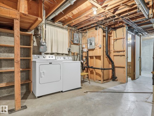 Basement laundry area featuring a washer and dryer pair, concrete flooring, exposed wood beam ceiling, built-in wooden shelving, and a utility sink visible through an open doorway - 13504 116 St Nw, Edmonton, AB - Indoor Photo Showing Laundry Room