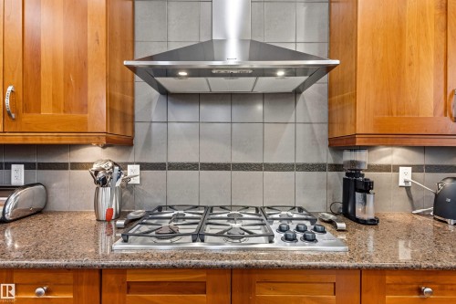 Stainless steel gas range with a coordinating range hood, granite countertops, wood cabinetry, and a tiled backsplash with a decorative accent strip - 1728 Rutherford Pt Point(E) Sw, Edmonton, AB - Indoor Photo Showing Kitchen With Upgraded Kitchen