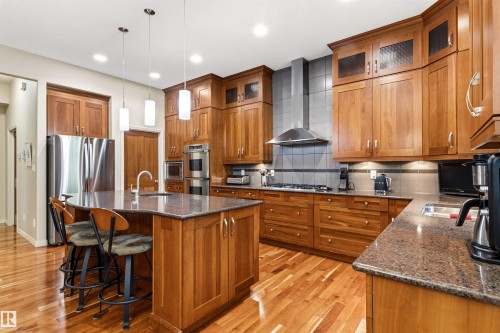 Kitchen featuring wood cabinetry, stainless steel appliances, a stone countertop island with seating, pendant lighting, and wood-finish flooring - 1728 Rutherford Pt Point(E) Sw, Edmonton, AB - Indoor Photo Showing Kitchen With Double Sink With Upgraded Kitchen