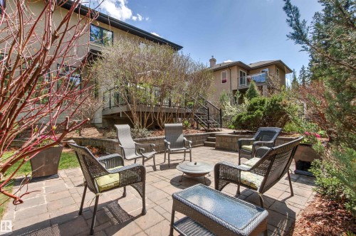 Paved stone patio featuring a built-in fire pit, surrounded by lush landscaping and a multi-level deck with black railings - 1728 Rutherford Pt Point(E) Sw, Edmonton, AB - Outdoor