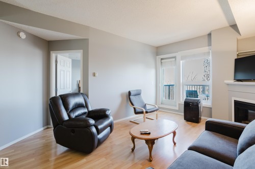 Living area featuring wood-finish flooring, a white fireplace with a dark insert, and a glass-paneled door opening to a balcony with dark railings - 508 11103 84 Avenue, Edmonton, AB - Indoor Photo Showing Living Room With Fireplace
