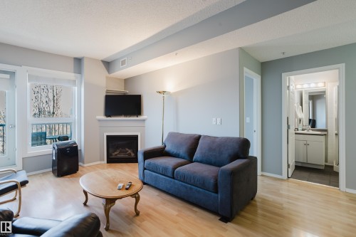 Living area featuring light wood-finish flooring, a corner fireplace with a white mantel, and a balcony with an outdoor railing - 508 11103 84 Avenue, Edmonton, AB - Indoor Photo Showing Living Room With Fireplace