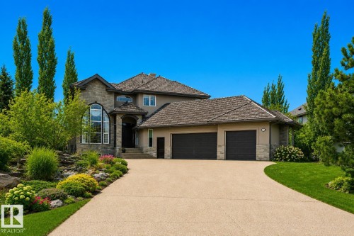 Stone and stucco exterior featuring a grand arched window, a multi-car garage, a concrete driveway, and established landscaping with mature trees and shrubs - 58 Riverstone Close, Rural Sturgeon County, AB - Outdoor With Facade