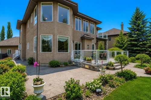 Stucco exterior featuring multiple large windows, a raised deck with glass panel railing, a lower stamped concrete patio, mature landscaping, and a manicured lawn - 58 Riverstone Close, Rural Sturgeon County, AB - Outdoor
