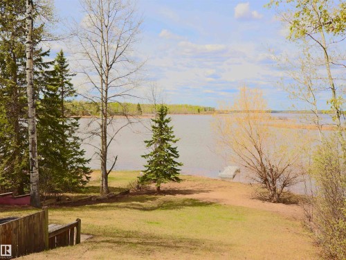 Waterfront property featuring a grassy bank, mature trees, a small wooden dock, and a distant shoreline - #26 5508 Twp Rd 593A, Rural Barrhead County, AB - Outdoor With View