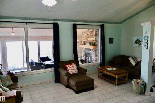 Spacious living area featuring light-colored tile flooring, two large windows, a ceiling fan, and a stone-faced fireplace with a wooden mantel - #26 5508 Twp Rd 593A, Rural Barrhead County, AB - Indoor Photo Showing Other Room