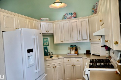 Kitchen featuring white cabinetry with gold-toned hardware, light-toned countertops, a white refrigerator with a dispenser, a white range, and a white range hood - #26 5508 Twp Rd 593A, Rural Barrhead County, AB - Indoor Photo Showing Kitchen
