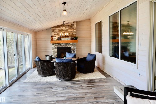 Spacious room featuring a stone fireplace with a wood mantel, wood-finish flooring, and white horizontal plank walls - #26 5508 Twp Rd 593A, Rural Barrhead County, AB - Indoor