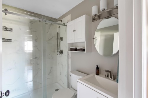 Modern bathroom featuring a glass-enclosed shower with white marble-look paneling, a white vanity with an integrated sink, a circular mirror, and a wall-mounted medicine cabinet - 803 12 Avenue, Cold Lake, AB - Indoor Photo Showing Bathroom