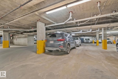 Underground parking garage featuring concrete flooring, exposed overhead piping, concrete support columns with yellow protective wraps, and fluorescent lighting - 206 103 Ambleside Drive, Edmonton, AB - Indoor Photo Showing Garage