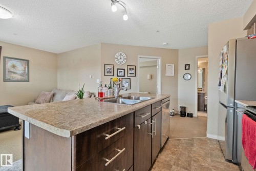 Kitchen island featuring a double basin sink, dark wood-finish cabinetry with silver-tone hardware, and a light-toned countertop - 206 103 Ambleside Drive, Edmonton, AB - Indoor Photo Showing Kitchen With Double Sink