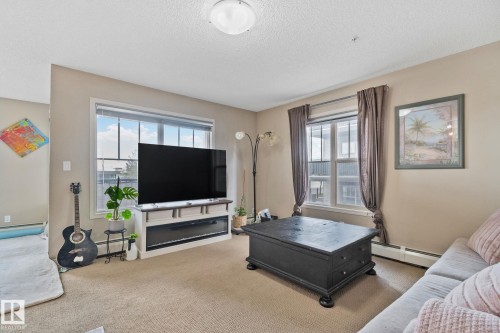 Carpeted living area featuring dual windows with blinds and drapes, a built-in media console with a linear fireplace, and neutral wall tones - 206 103 Ambleside Drive, Edmonton, AB - Indoor
