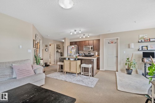 Open-concept living space featuring neutral carpet and paint, recessed ceiling lighting, and a kitchen with dark wood-finish cabinetry, stainless steel appliances, and a breakfast bar - 206 103 Ambleside Drive, Edmonton, AB - Indoor