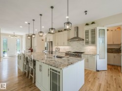 Spacious kitchen featuring a large island with a sink, pendant lighting, stainless steel appliances, light-toned cabinetry, and wood-finish flooring - 