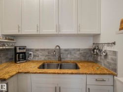 Kitchen featuring white shaker-style cabinetry, a double stainless steel sink with a gooseneck faucet, light granite countertops, and a gray subway tile backsplash - 