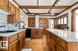 Spacious kitchen featuring extensive wood cabinetry, light-toned countertops, a black double-basin sink, wood-finish flooring, and decorative ceiling beams with track lighting - 