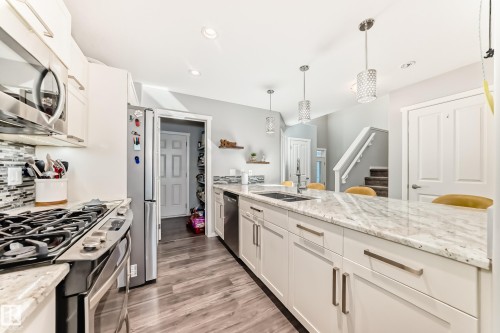 Open-concept kitchen featuring white cabinetry, stainless steel appliances, wood-finish flooring, and a large central island with a light-toned countertop and pendant lighting - 267 Sturtz Bend, Leduc, AB - Indoor Photo Showing Kitchen With Double Sink With Upgraded Kitchen