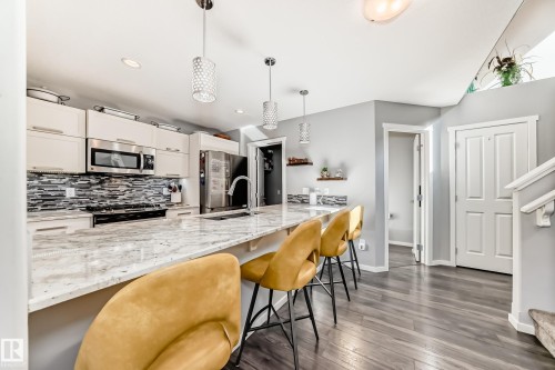 Kitchen featuring an expansive stone countertop island with pendant lighting, stainless steel appliances, a mosaic tile backsplash, wood-finish flooring, and light-toned cabinetry - 267 Sturtz Bend, Leduc, AB - Indoor