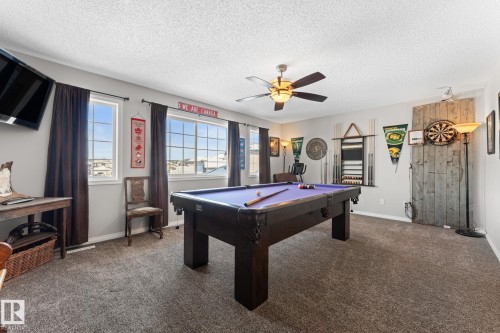 Spacious recreation room featuring a built-in pool table, broad windows, textured carpeting, a ceiling fan with integrated lighting, and a prominent wood-panel accent wall - 112 Lakeland Point(E), Beaumont, AB - Indoor Photo Showing Other Room