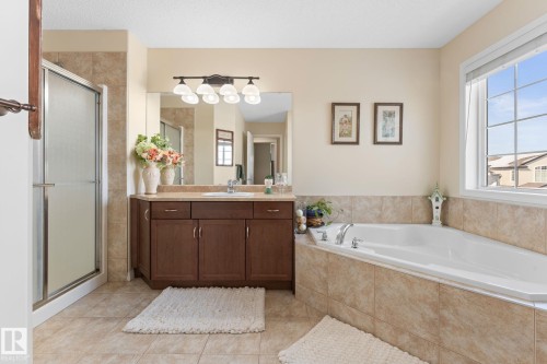 Bathroom featuring a corner soaking tub, a tiled shower with a glass door, a single vanity with dark wood cabinetry and a light-colored countertop, a large mirror, and tiled flooring - 112 Lakeland Point(E), Beaumont, AB - Indoor Photo Showing Bathroom