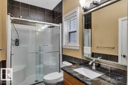 Bathroom featuring a glass-enclosed shower with integrated seating, a dark tiled shower surround, a vanity with a dark stone countertop and integrated sink, a large framed mirror, and a window with blinds - 
