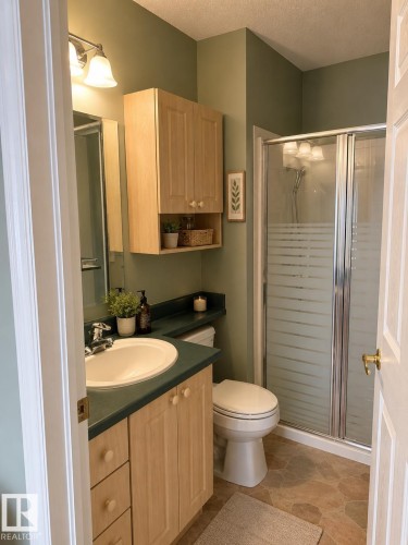 Full bathroom featuring a single vanity with wood-finish cabinetry and a green countertop, a white toilet, and a glass-enclosed shower with frosted panels - 219 9535 176 Street, Edmonton, AB - Indoor Photo Showing Bathroom