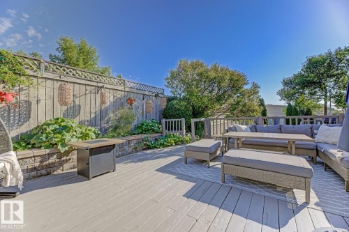 Spacious outdoor deck featuring light-toned wood-finish planks, a perimeter wooden fence with lattice detailing, and a stone retaining wall supporting abundant green foliage - 3208 135A Avenue, Edmonton, AB - Outdoor With Deck Patio Veranda With Exterior